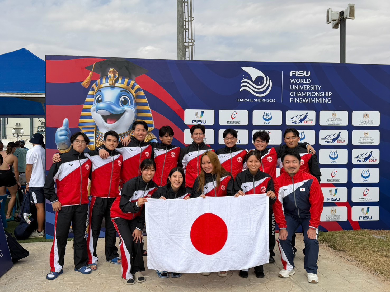 2026 FISUフィンスイミング世界大学選手権の学生日本代表。大会マスコットと大会ロゴのボードの前で日の丸を掲げた集合写真